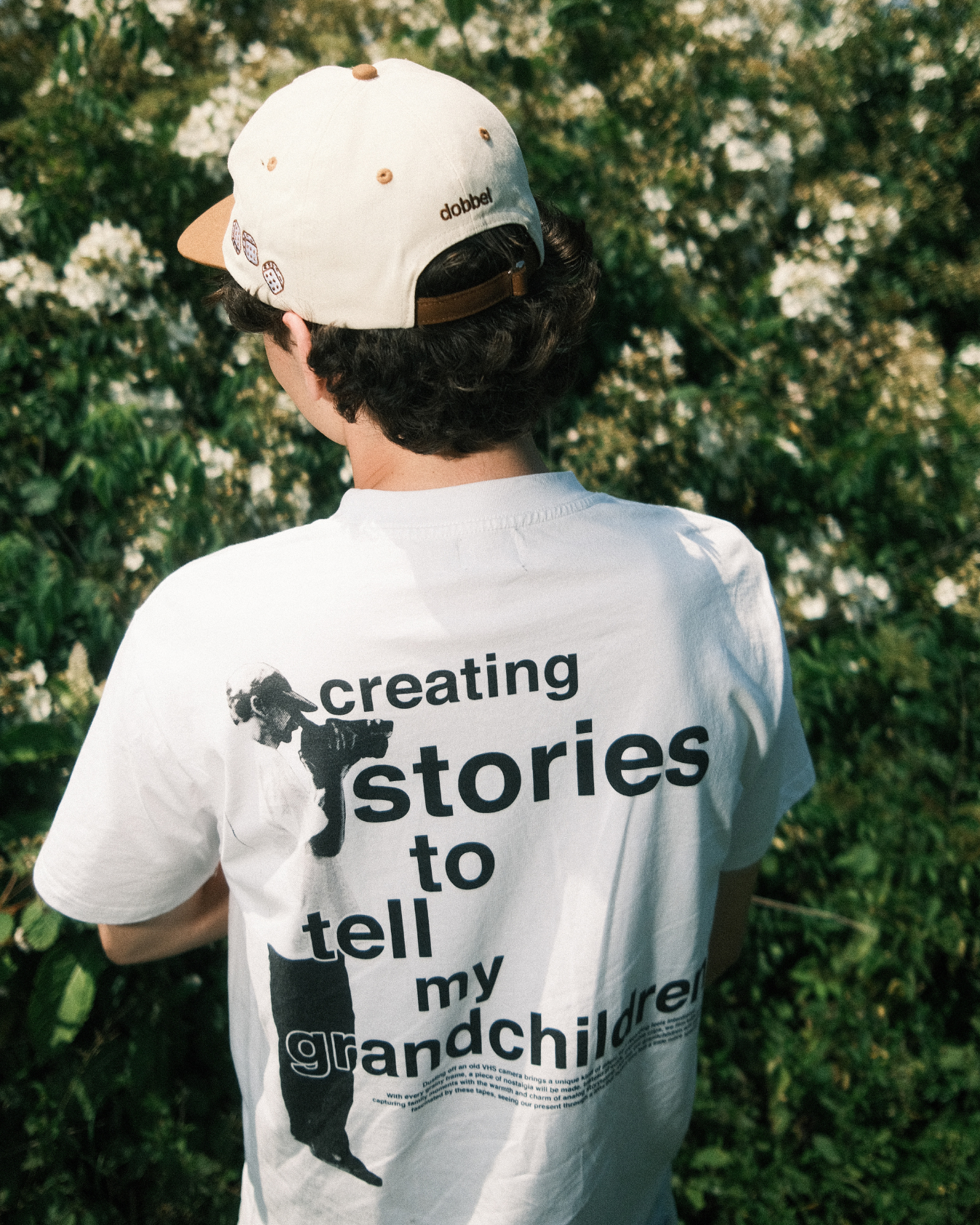 Person wearing a white dobbel vhs t-shirt with text and a cap, standing in front of green foliage.