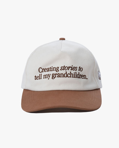 White cap with brown brim and text 'Creating stories to tell my grandchildren' with 'dobbel' on the back of the cap
