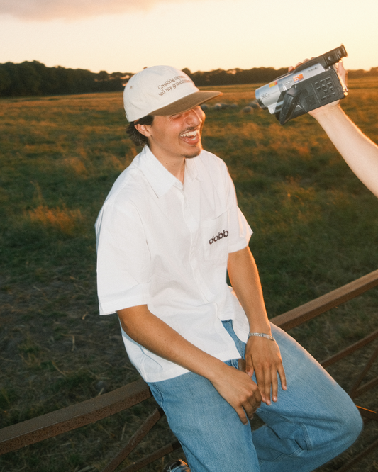 Man in a white dobbel creating stories to tell my grandchildren shirt and dobbel legacy cap holding a vhs camera in a field