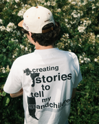 Person wearing a white dobbel vhs t-shirt with text and a cap, standing in front of green foliage.